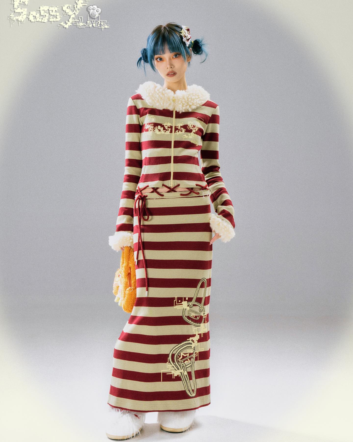 the image shows a fulllength studio shot of a young asian woman wearing a long striped dress. the dress is primarily red and beige with horizontal stripes and features a faux fur collar. it also has a graphic design printed on it. she is accessorized with a small fluffy bag and fluffy white boots. the model has blue hair styled in space buns and is posed against a light grey background.  the image appears to be a fashion photograph showcasing the dress from a clothing brand labeled "sassy flower".