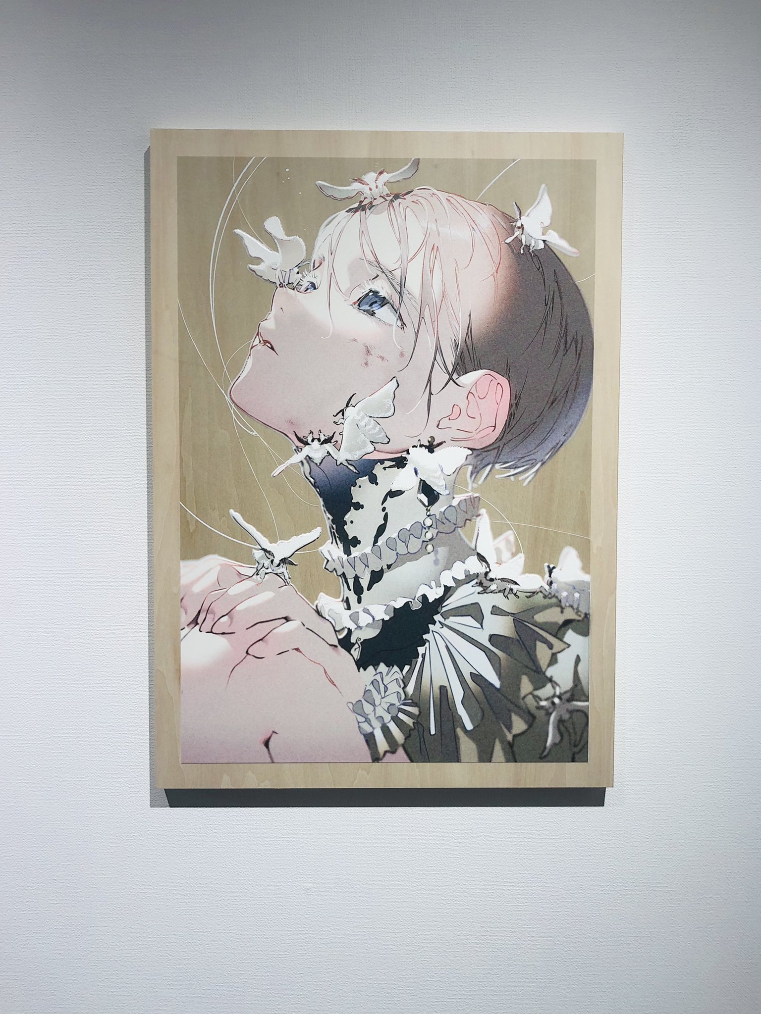 the image showcases a digital illustration printed on a wooden board and displayed in a gallery setting. the illustration depicts a young man with short, stylized hair, his head tilted back and eyes closed. several white butterflies rest on his neck and chest, their delicate forms mirroring the softness of his expression. he wears an intricate, ruffled collar, hinting at a sense of elegance and fragility. the background features a subtle circular pattern in pale gold, adding to the ethereal ambiance.