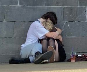 a couple sitting on the ground in front of a concrete wall. they are hugging, with the boy's arm around the girl's shoulders. the boy is wearing a white shirt and blue jeans, and the girl is wearing a black skirt and black stockings.