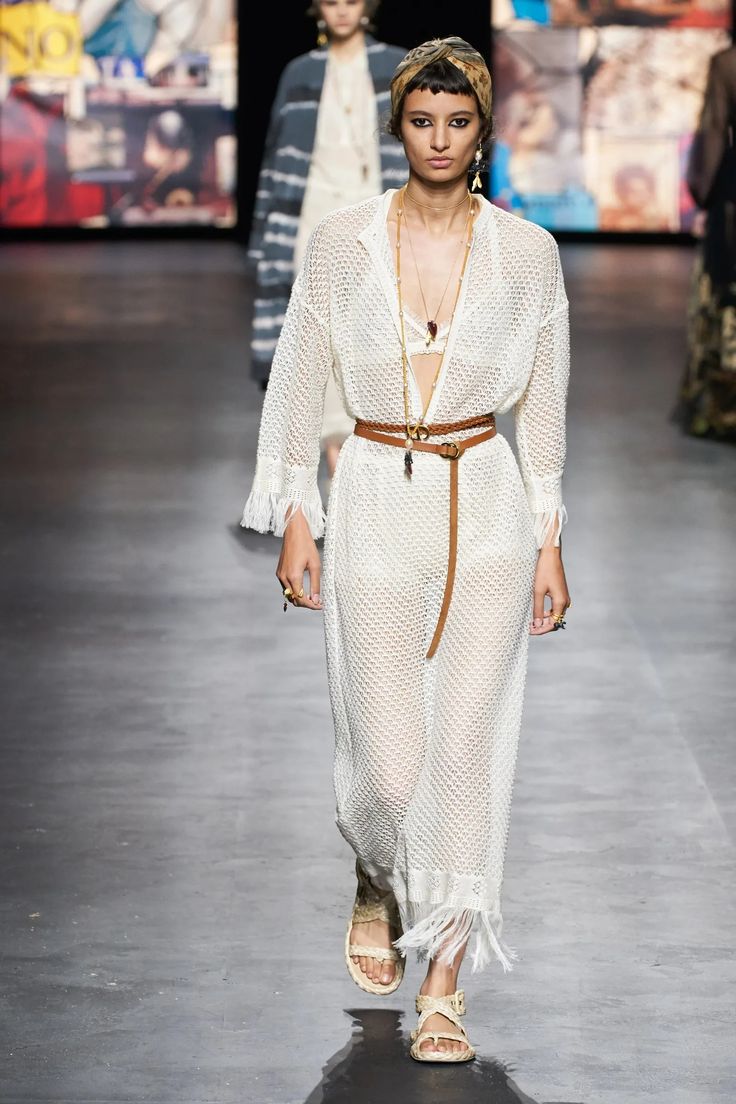 a female model in a white crochet dress walks down the runway. the dress is belted at the waist with a brown leather belt and she is wearing a pair of tan sandals. she has her hair tied up in a headband.