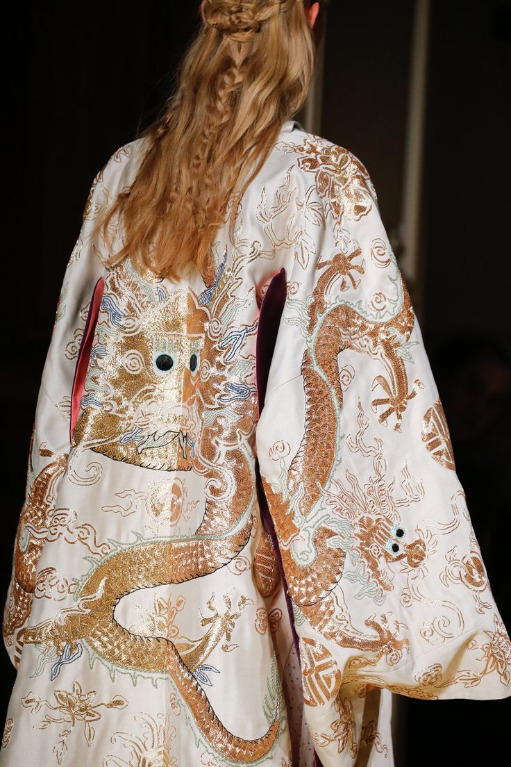the image shows the back of a model wearing a long, elegant creamcolored robe or kimono. the garment is heavily embroidered with a large gold dragon design, intricately detailed and flowing down the length of the robe. the embroidery also features other floral and symbolic elements. the model has long blonde hair styled in a braid. the background is dark and out of focus, highlighting the garment's detail and texture. the overall impression is one of high fashion, luxury, and intricate craftsmanship.