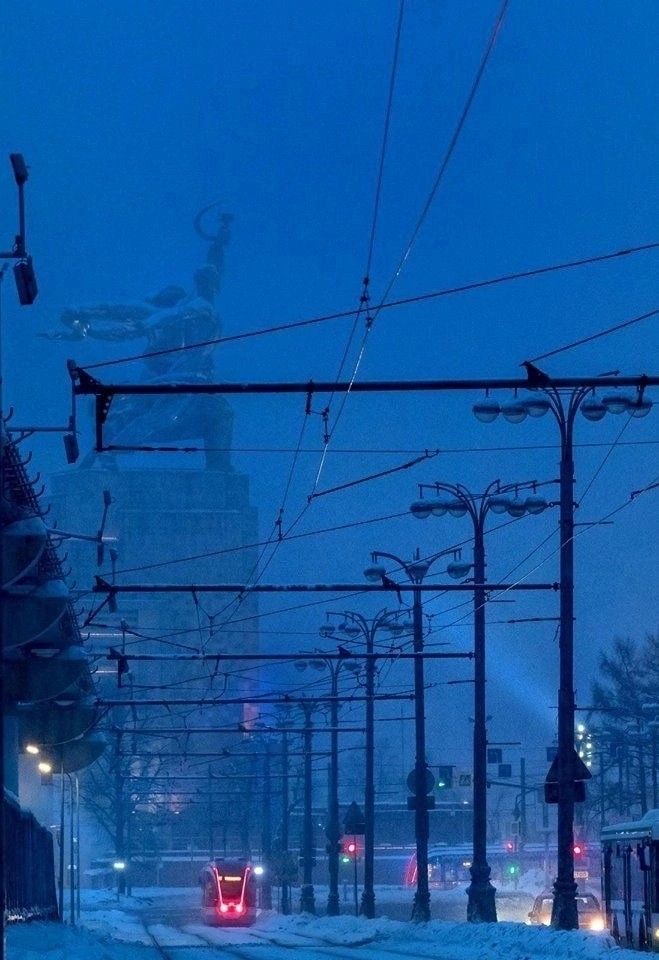the image shows a snowcovered city street at twilight, with a tram traveling down the middle.  the street is lined with tall streetlights and power lines crisscrossing overhead. in the background, the imposing motherland monument is visible, partially obscured by the snow and atmospheric haze. the overall color palette is a deep, cool blue, enhancing the wintery atmosphere.  the scene is peaceful and serene, capturing a quiet moment in the city.