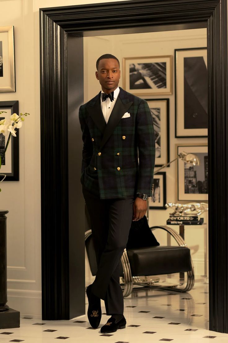 a full shot of a male model standing in a doorway, dressed in a luxurious ralph lauren doublebreasted green plaid suit. the suit is paired with a crisp white shirt, a black bow tie, and black velvet slippers. the model's sophisticated posture and the opulent interior setting contribute to the overall image of refined elegance and highend fashion. the background showcases framed artwork and a stylish chrome chair, further emphasizing the luxurious ambiance.