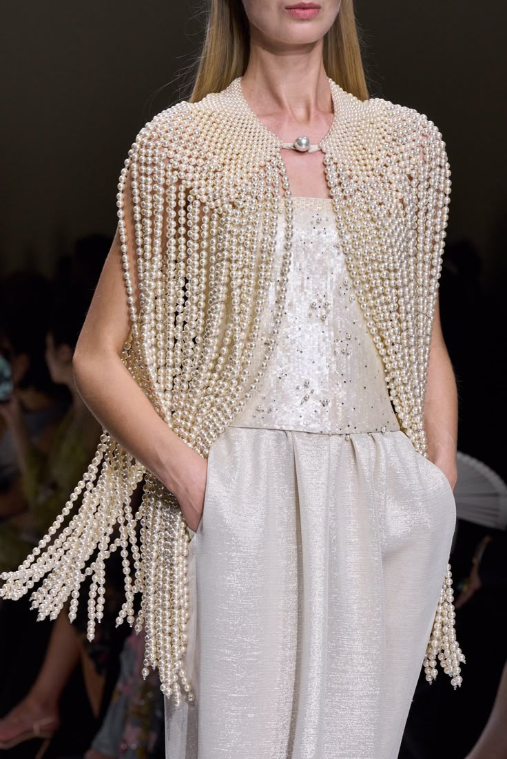 a woman is shown wearing a pearl cape with pearl fringe over a sequined top and dress pants. the outfit is on a runway.