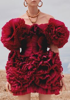 a woman is wearing a red ruffle dress that looks like a flower. the dress is very stylish and elegant. she is posing in a stylish manner. she has a necklace, earrings and a ring on her finger which adds to the overall glamorous look.