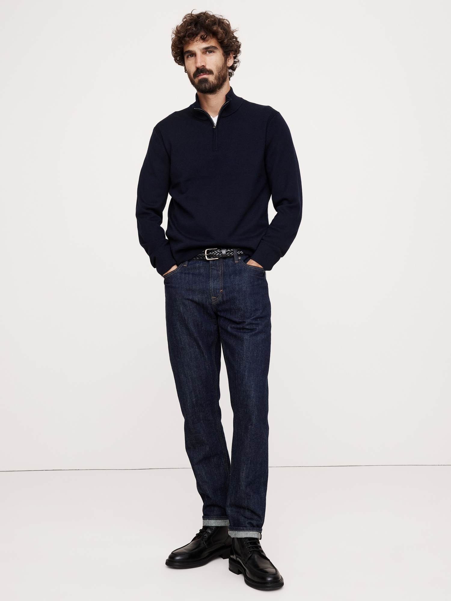 a full shot of a man standing in a studio setting with a solid white background. he is wearing a navy blue quarterzip pullover, dark wash jeans, a leather belt, and black leather boots. the man has curly hair and a beard. he has his hands in his pockets.