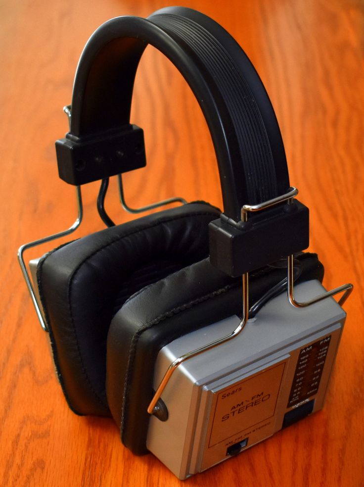 a vintage am/fm radio and headphone set sits on a wooden table. the headphones are black with leather ear cups and a padded headband. the radio unit is silver and has a series of knobs and buttons, as well as a display that shows the station frequency and other information. the radio is a portable model with a headphone jack, so it can be enjoyed in private or in quiet environments.  the headphones and radio are an example of 1970s1980s technology.