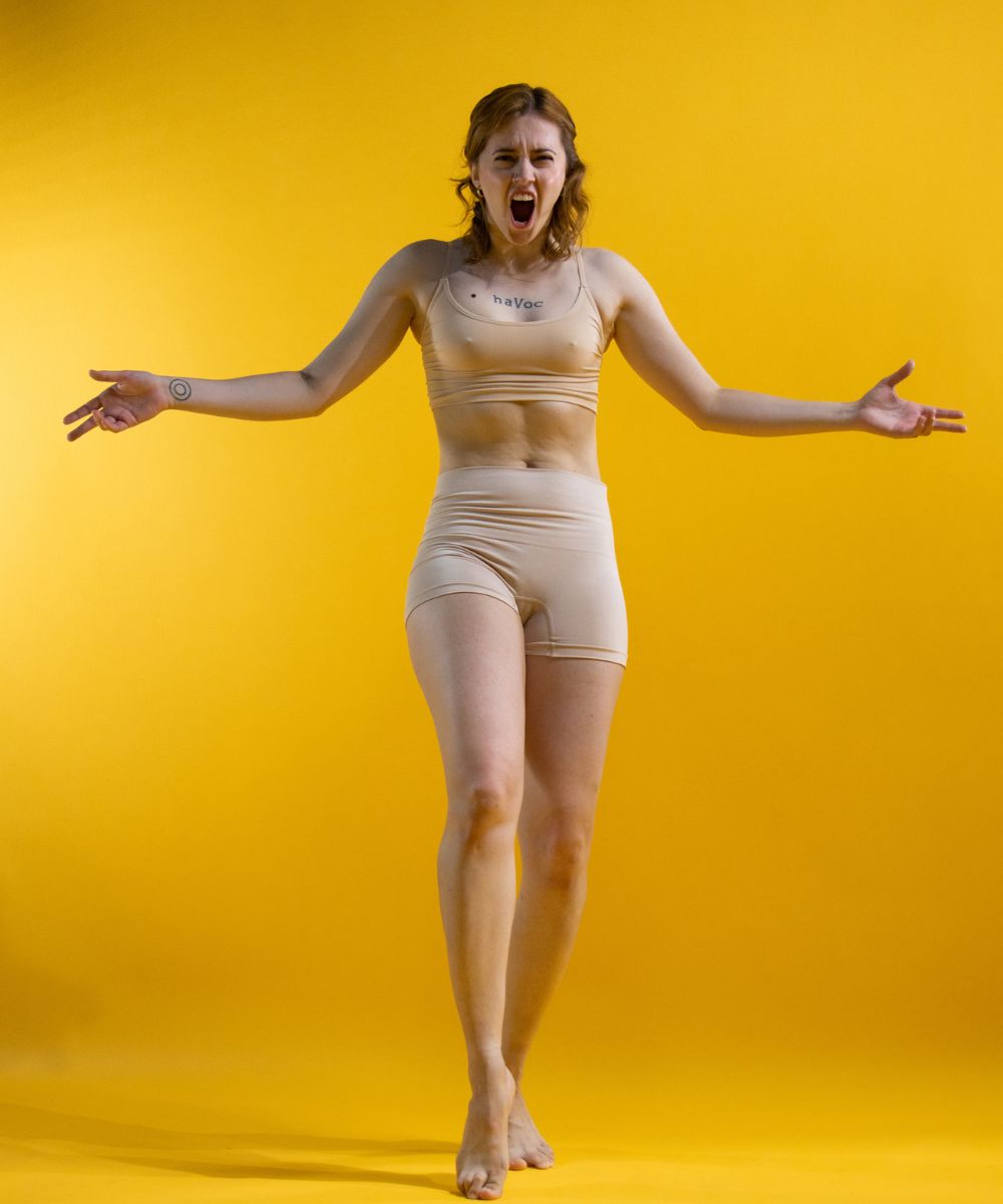 the image shows a young, slender woman with light skin standing against a bright yellow background.  she's wearing a light beige sports bra and shorts and is shown in a fullbody shot.  her arms are outstretched, and her mouth is open in a loud shout or scream, conveying a strong emotional expression of anger or frustration. she has a small tattoo that says "havoc" on her chest. the overall style is minimalist and the focus is on her emotional display.