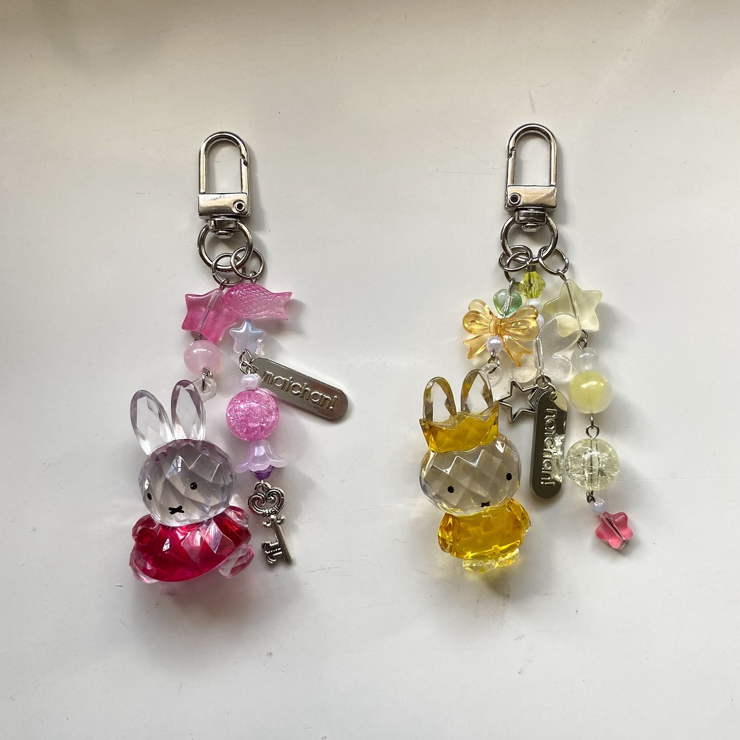 two keychains are laying next to each other on a white surface. each keychain has a silver clip at the top attached to a beaded chain. one keychain features a clear crystal bunny wearing a red dress, adorned with pink and white beads, a pink star, and a silver tag with "maichan" printed on it. the other keychain has a clear crystal bunny wearing a yellow crown and dress, accompanied by yellow and white beads, a bow, a star, and a silver tag with "notchan" written on it.