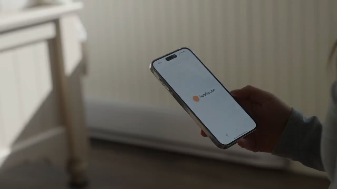 a woman is holding an iphone in her hand. the phone is open to the home screen of an app called "headspace." the background is a blurred out view of a room with a white wall and a white dresser. the room is lit by natural light coming from a window.