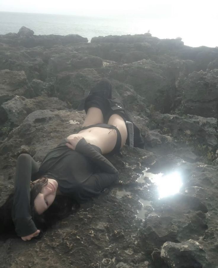 a woman is lying on her back among jagged rocks on a coast, with the ocean and a cloudy sky in the background. she is wearing a dark longsleeved shirt and a swimsuit. a small pool of water reflects the sunlight.