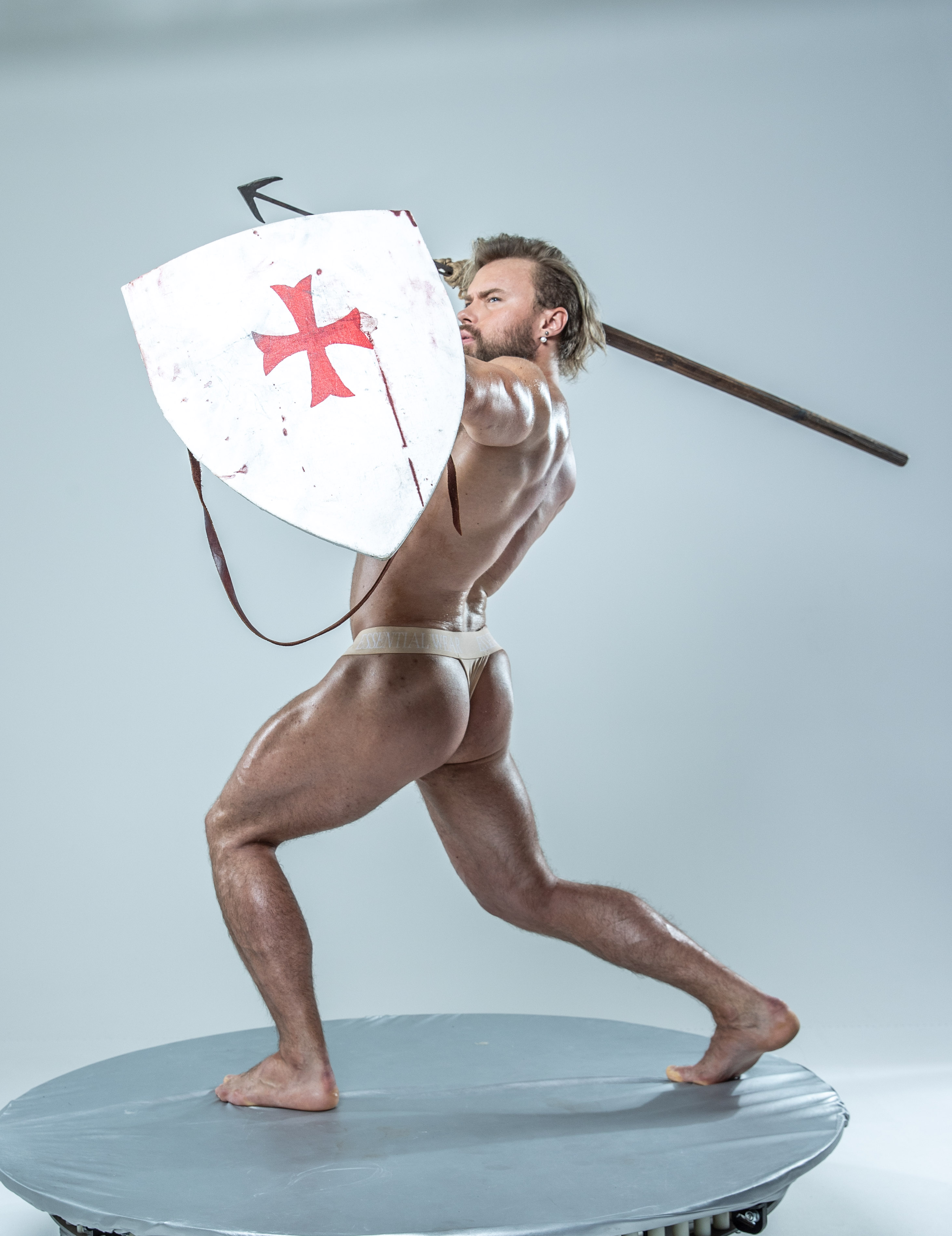 a muscular man, wearing only underwear, poses dynamically on a circular platform against a plain grey background. he brandishes a medievalstyle spear and holds a weathered shield adorned with a red cross, evoking the image of a historical warrior. the artificial lighting highlights his physique and the weaponry.