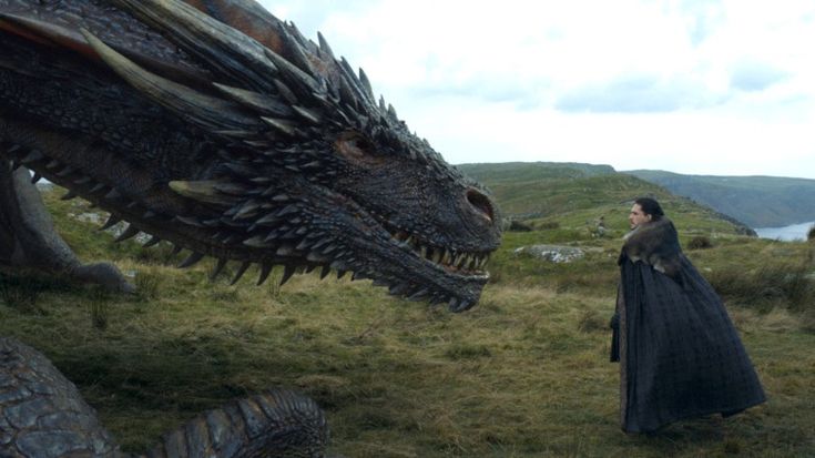 a still image from the television series game of thrones depicts jon snow (kit harington) standing before drogon, one of daenerys targaryen's dragons.  drogon is a massive, darkcolored dragon, partially visible in the foreground. jon snow is wearing his dark cloak with fur trim, his back to the camera as he looks towards the dragon.  the setting is a rugged, coastal landscape with hills and a glimpse of the ocean in the background. the scene evokes a sense of power, mystery, and the epic scope of the show.