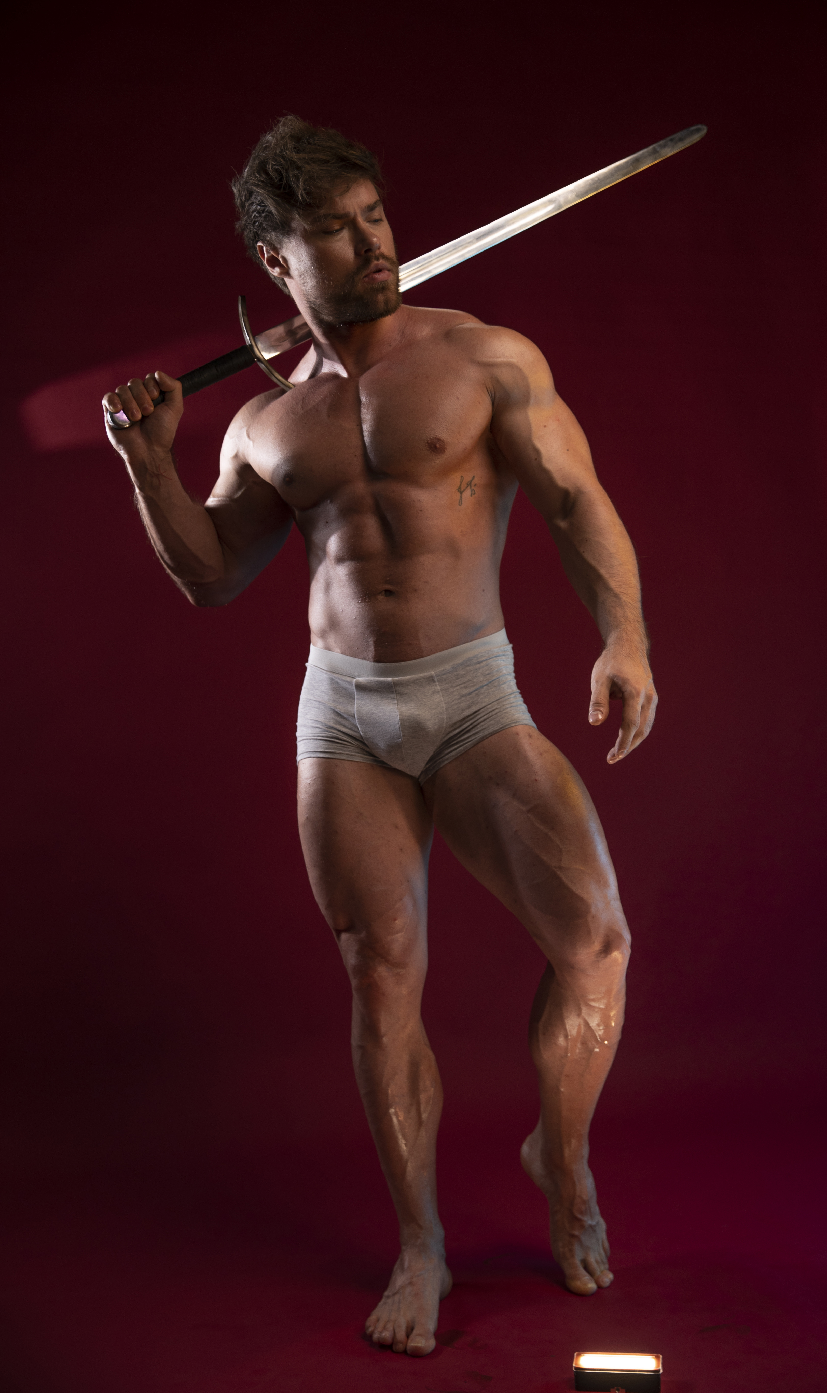 a muscular man is posing barechested with a sword resting on his shoulder. he is wearing underwear and is standing against a red background. the lighting highlights his physique.