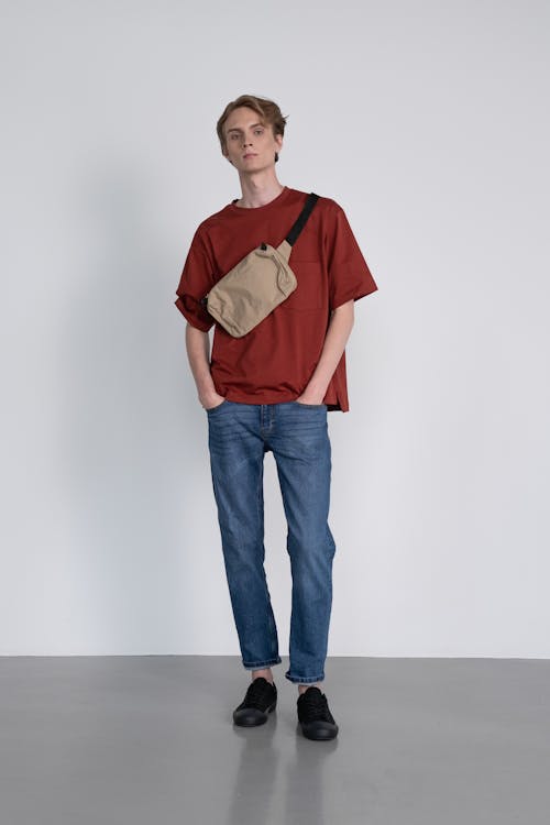 a fulllength studio shot of a male model standing with his hands in his pockets, wearing casual attire. he is dressed in a reddishbrown tshirt, blue jeans, and black sneakers. he is also wearing a light brown crossbody bag over his shoulder. the background is a plain white wall and a gray floor. the model is looking directly at the camera.
