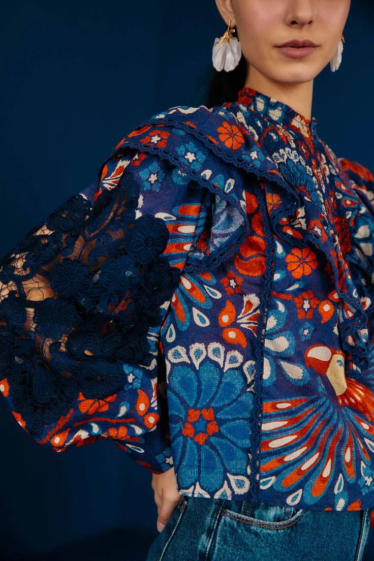 closeup view of a woman wearing a vibrant blue and orange floral blouse with navy lace sleeves and ruffles. the blouse has a high neck and long sleeves. the woman is also wearing denim jeans and large statement earrings. the backdrop is a deep blue. the image focuses on the intricate details of the blouse's pattern and texture. the overall style is bohemian and slightly retro.