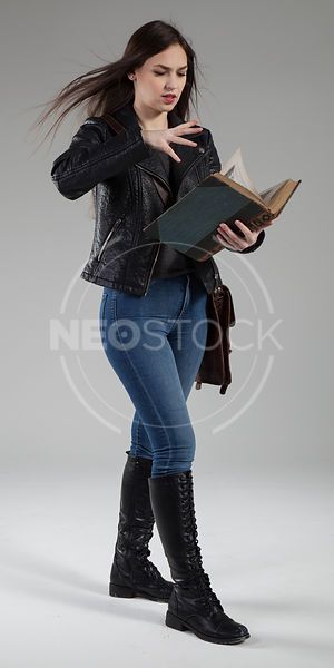 a woman in a leather jacket and jeans stands in a studio, looking down at a book in her hand. she's wearing high heeled boots and has long, dark hair.  she's looking at the book with a serious expression on her face.