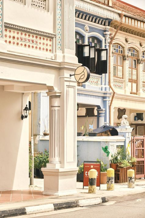 sunlit street scene in singapore featuring a row of beautifully preserved shophouses. the focus is on a creamcolored shophouse with a cafe sign,  featuring classical architectural details like pillars and ornate tilework. the neighboring shophouses are painted in pastel shades of blue and beige, showcasing a mix of architectural styles. plants in pots add a touch of greenery to the scene. the overall impression is one of charming, historic urban architecture.