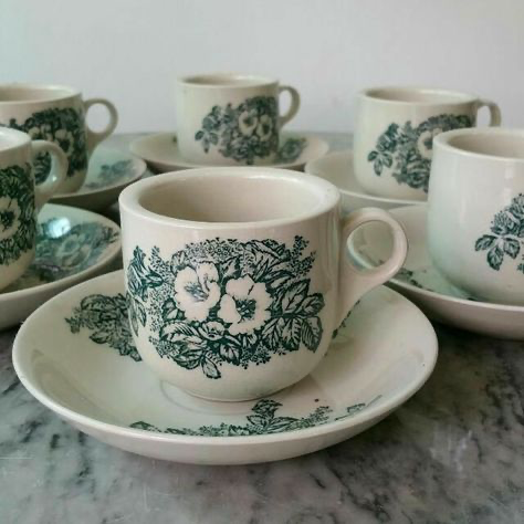 close up image of a table with a vintage floral tea set in green and white on a white marble surface. the image shows 6 teacups and saucers, all with a floral pattern.  the teacups are filled with tea and the saucers are placed neatly on the table. the background is blurred and the focus is on the tea set.