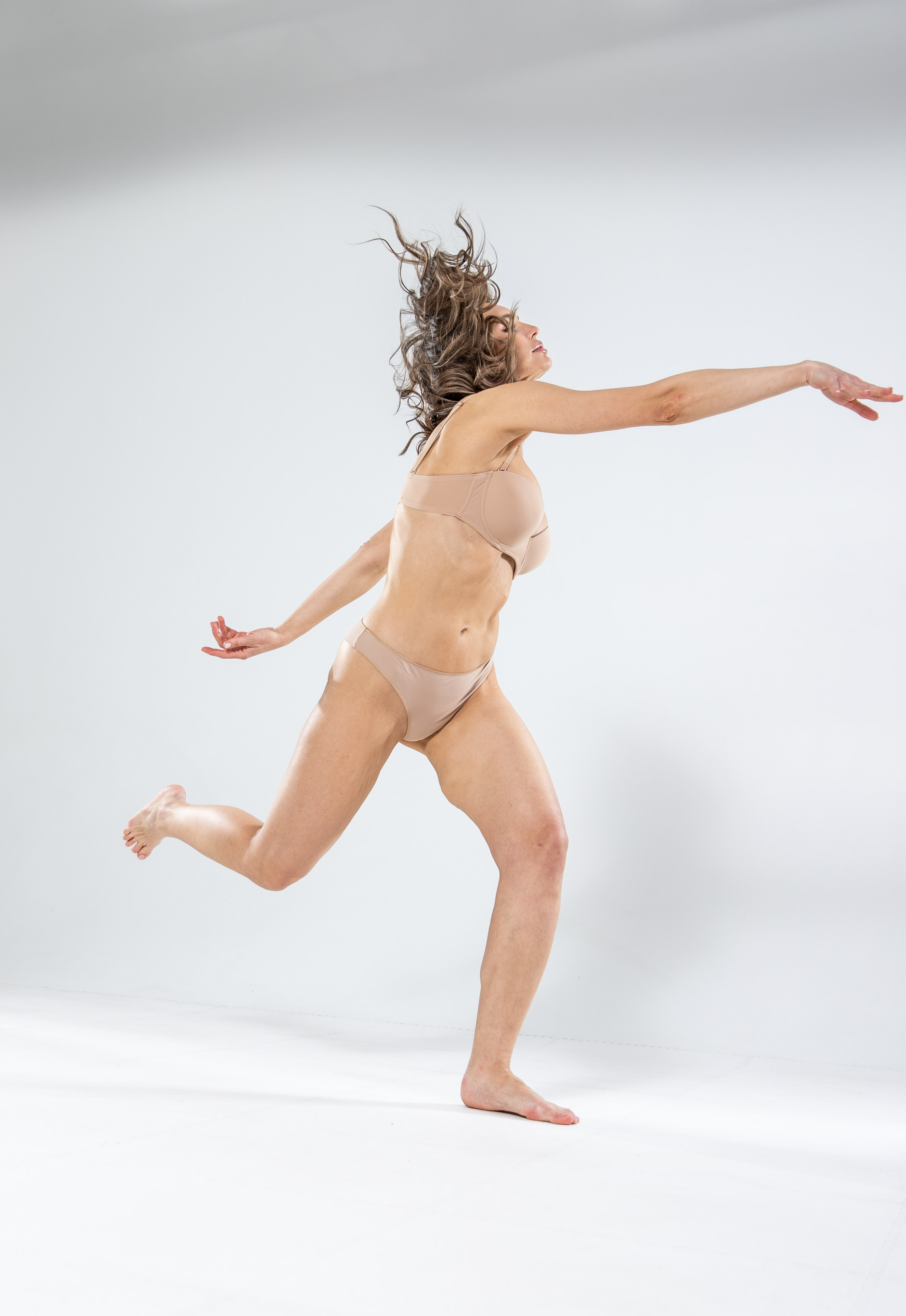a woman is captured in a dynamic pose against a white background. she appears to be dancing or running, with her curly hair flowing behind her. she is wearing nudecolored underwear and is barefoot. the studio lighting highlights her skin and the contours of her body. the shot is a full view, showcasing her entire figure in motion.