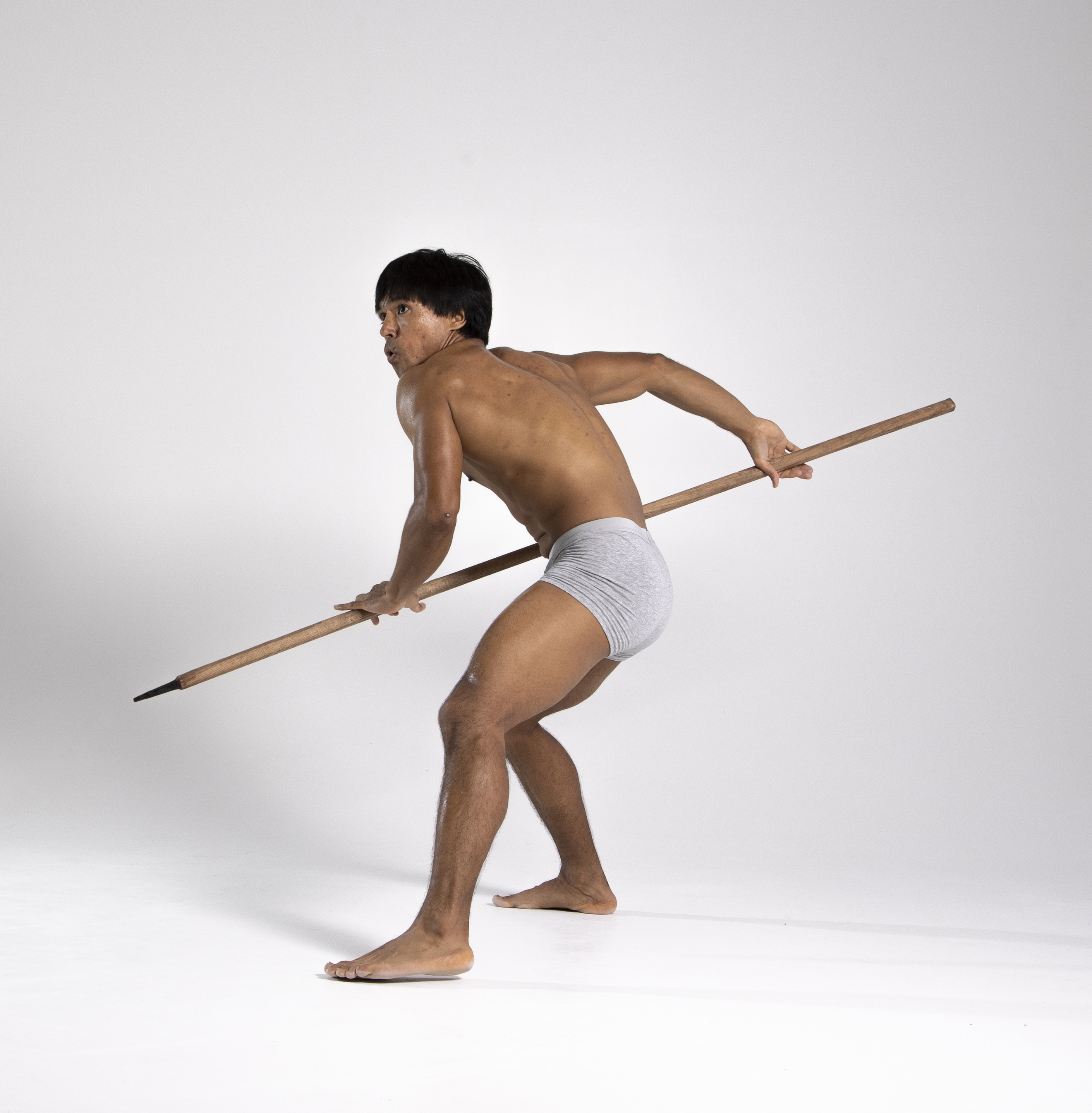 a man, possibly indigenous, is shown in a studio shot, holding a spear. he is wearing a loincloth and has bare feet. he appears to be a warrior, possibly in a defensive stance, with the spear as his weapon. he is seen from a rear view.