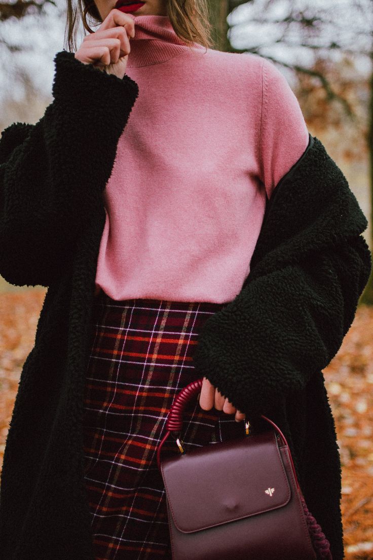 a closeup shot of a woman wearing a stylish autumn outfit. she's wearing a pink turtleneck sweater, a red and black plaid skirt, and a black teddy coat.  she's holding a burgundy handbag. the background is blurred but shows autumnal leaves on the ground. the woman's expression is slightly coy, with her hand near her mouth. the overall aesthetic is chic and autumnal.
