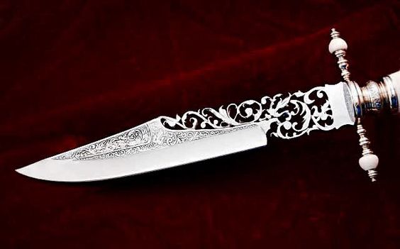 the image showcases a closeup of an exquisitely crafted dagger with intricate engravings covering the blade and handle. the blade, seemingly made of polished steel, reflects light, highlighting the detailed artistry. the handle, possibly made of silver, features delicate, flowing patterns and embellishments, indicating fine craftsmanship. the dagger rests on a rich, dark red velvet background, enhancing its elegance and beauty.