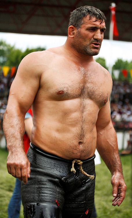 a shirtless man, a turkish oil wrestler, poses for the camera. he is very muscular and has a powerful build.  he wears a traditional pair of black pants with a rope tie. he is standing in a field and has a serious expression on his face.