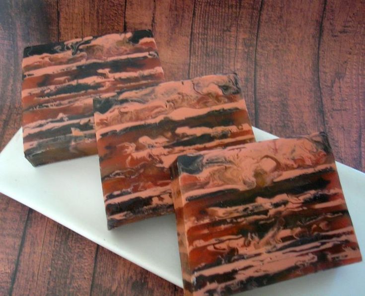 three bars of artisan soap with a layered orange, brown and black swirl design are displayed on a white rectangular plate, with the background of a dark brown woodgrain surface.