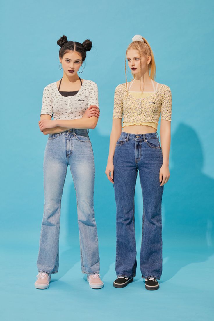two young women are standing sidebyside against a light blue background. they are both wearing jeans and shortsleeved tops. the woman on the left is wearing light wash bellbottom jeans and a white floral top. the woman on the right is wearing dark wash bellbottom jeans and a light yellow floral crop top. they both have their hair styled in updos. they are both wearing sneakers.