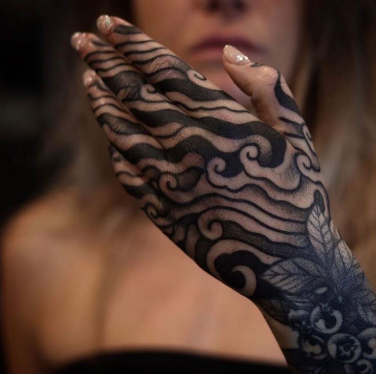 the image shows a woman's hand with a large, intricate tattoo design. the tattoo features a wave pattern of black ink across the fingers and hand, with swirling, curved lines that resemble water or clouds. the design continues down the arm, incorporating a floral element with leaves and possibly berries. the woman has nail polish on her fingernails. her shoulder and arm are visible, and the overall effect is striking and artistic.