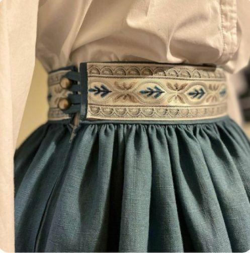 loseup view of a woman's teal skirt with a unique embroidered waistband. the skirt is made of a linenlike material, and it is gathered at the waist. the waistband is decorated with an intricate embroidered design in shades of cream, beige, and light blue. a white blouse is visible underneath the skirt, and the overall style is reminiscent of vintage or folk costume. the skirt appears to be highwaisted and the buttons on the back waistband are visible. the focus is on the texture and detail of the fabric and embroidery.