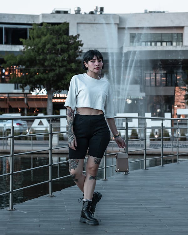 a young woman with short black hair and tattoos stands on a walkway in a modern city setting. she's wearing a white crop top, black biker shorts, combat boots, and a beige crossbody bag. a fountain is visible in the background. the overall style is urban and trendy.  she looks directly at the camera with a confident expression.