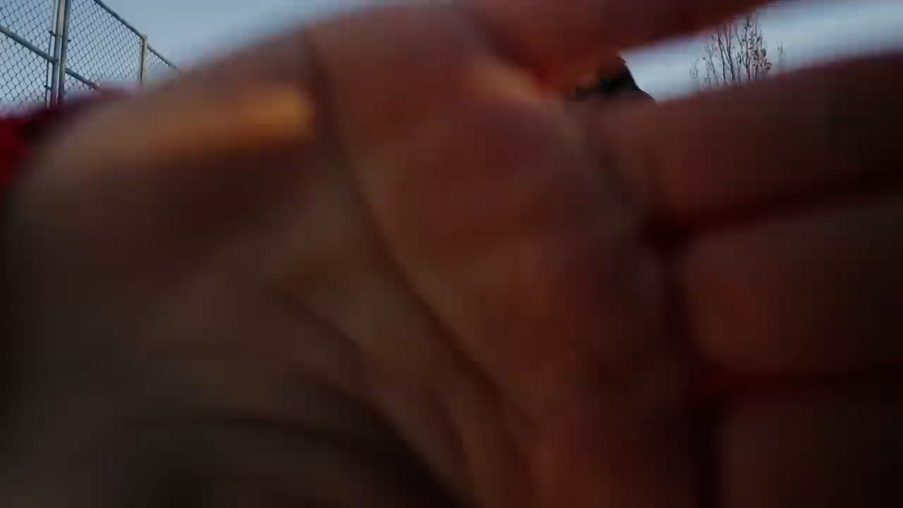 a closeup shot of a hand partially obscuring the view. the hand is large in the frame, creating a sense of immediacy and a firstperson perspective. the fingers are slightly spread, revealing a glimpse of a person in the background, along with a chainlink fence and a cloudy sky. the overall lighting suggests late afternoon, with soft, diffused light casting a cool tone over the scene.