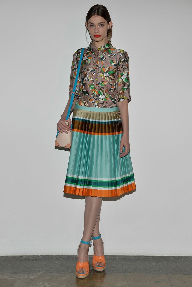 a full shot of a female model posing against a white background. she is wearing a floral print shirt paired with a teal pleated skirt featuring orange and brown stripes. the model also wears a light peachcolored handbag with a light blue strap slung across her body and bright orange sandals with teal straps.  the overall style is a vibrant summer look.