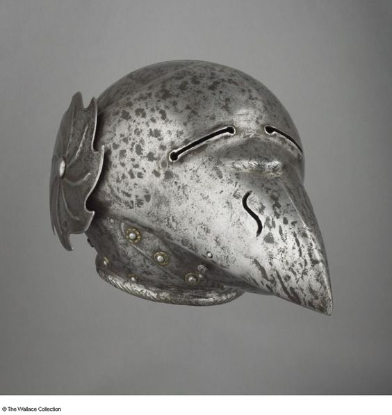 a closeup view of an antique medieval helmet, crafted from steel. its design features a distinctive birdlike beak and intricate metalwork. the helmet is displayed against a neutral gray background, suggesting it's likely part of a museum collection.