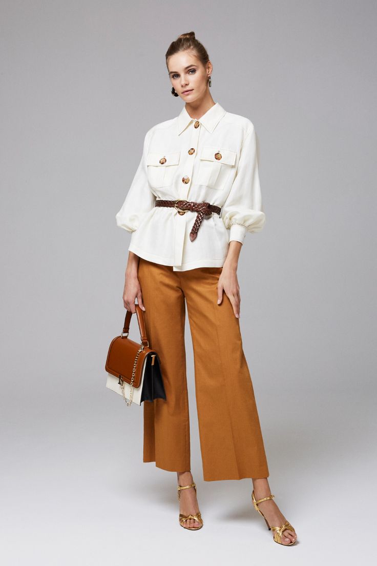 a fullbody studio shot of a woman posing against a light grey background. she's wearing a creamcolored collared shirt with large buttons, brown wideleg pants, and gold sandals. she's accessorized with a brown braided belt, a brown and cream handbag, and gold earrings. her hair is pulled back in a bun. the overall style is elegant and chic.
