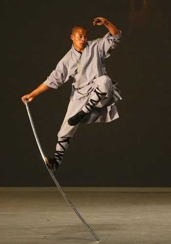 a shaolin monk in white robes demonstrates his martial arts skills by balancing on a sword in midair. the monk's focused expression and dynamic pose convey the power and discipline inherent in his practice.