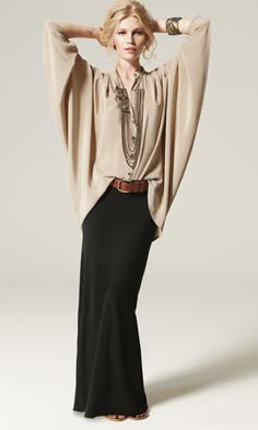 a woman is wearing a longsleeved beige blouse with a black maxi skirt and a brown belt. she is also wearing a necklace and a bracelet. she is standing in a studio, and the background is white. she is looking at the camera. the woman is wearing a casual but elegant outfit.