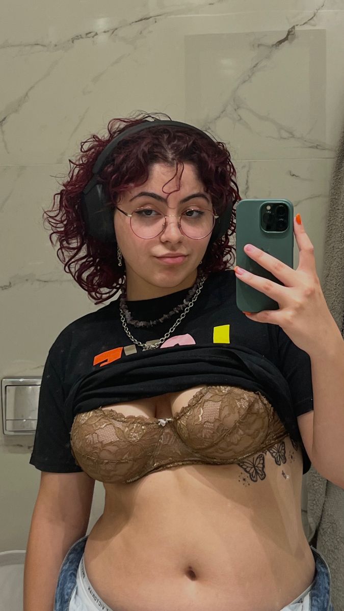 a selfie of a woman with curly red hair, wearing glasses and headphones. she is wearing a black tshirt pulled up to expose her stomach and a brown lace bra. she has a tattoo on her side and is wearing jewelry. she is holding a green phone.