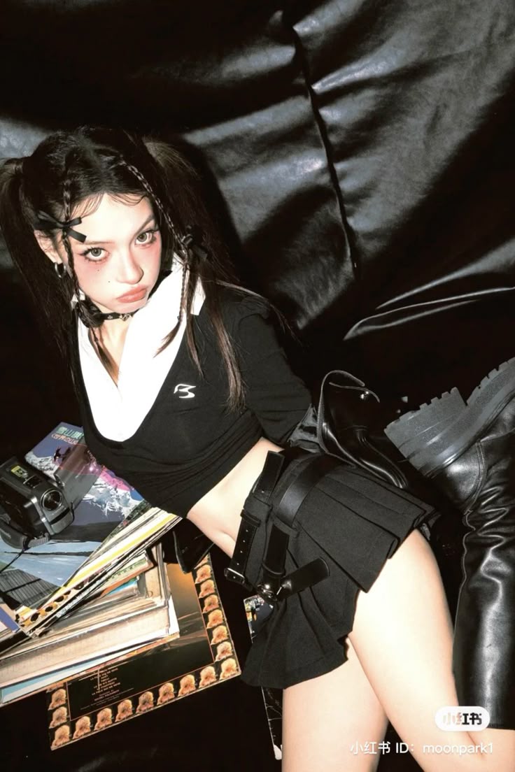 a woman with pigtails and braids is posing in a punkinspired outfit. she is wearing a black crop top with a white collar, a black pleated skirt with a belt, and black boots. she is wearing makeup that includes blush and eyeliner. she is surrounded by records and a camera. she is wearing a choker.