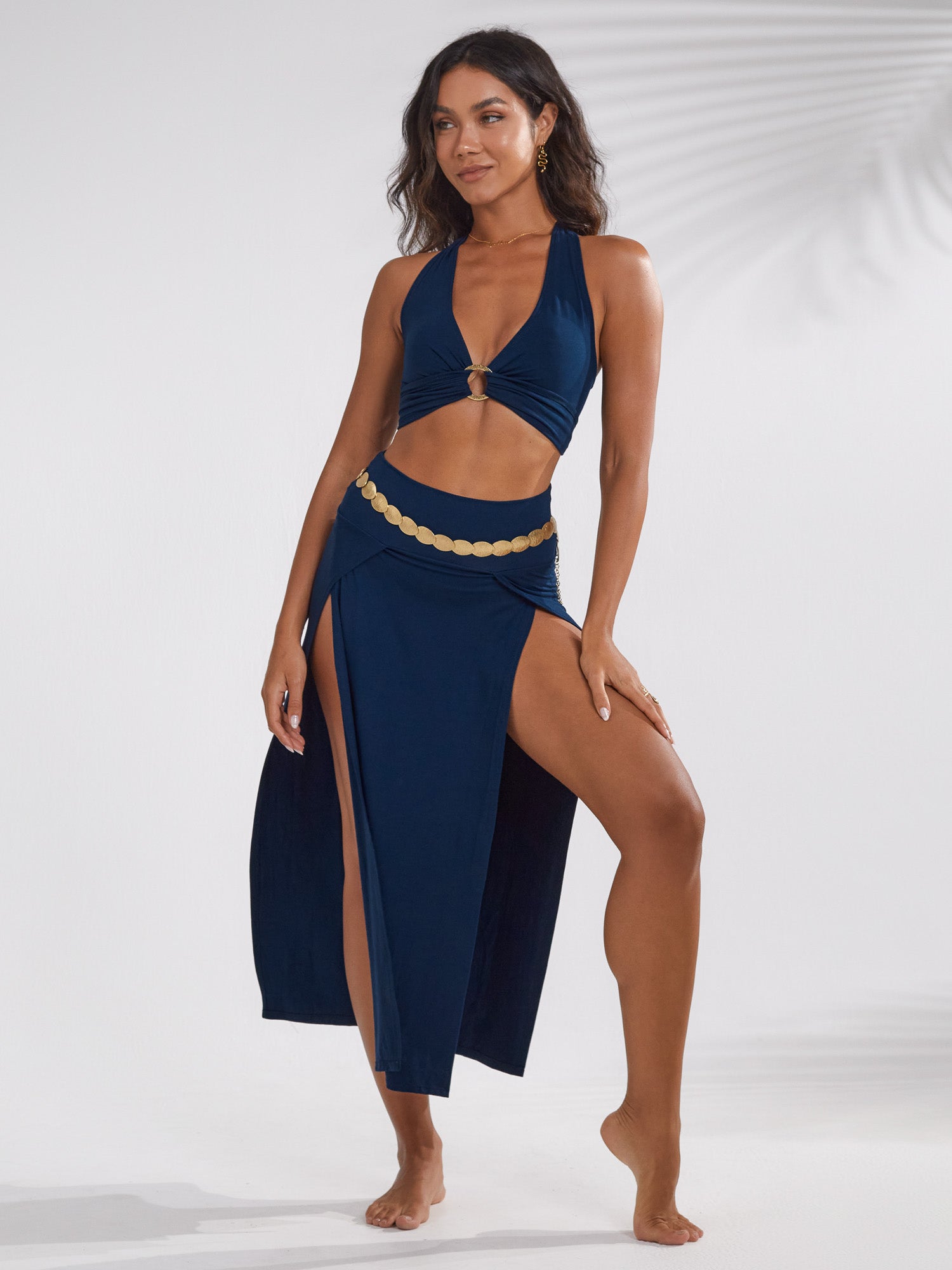 a fulllength shot of a beautiful woman model posing in a navy blue halter top and matching maxi skirt with a gold coin belt. the skirt features a high slit on each side. she's standing on a lightcolored background, with her hair styled elegantly. the overall style is chic and beachready.
