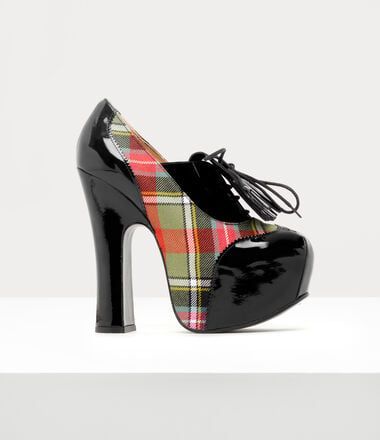 a single platform shoe is shown. the shoe has a plaid tartan pattern on its sides, black patent leather around the toe and the heel, as well as a black tie with tassels on the front.