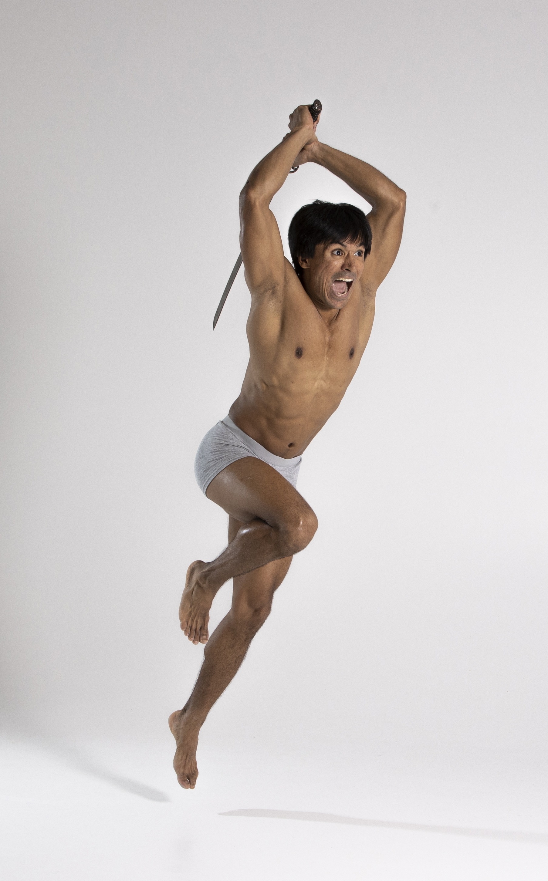 a shirtless asian male is jumping and screaming while holding a sword. he is wearing light gray underwear and is barefoot. the background is a light gray studio setting. he is holding the sword above his head with both hands, ready to strike.