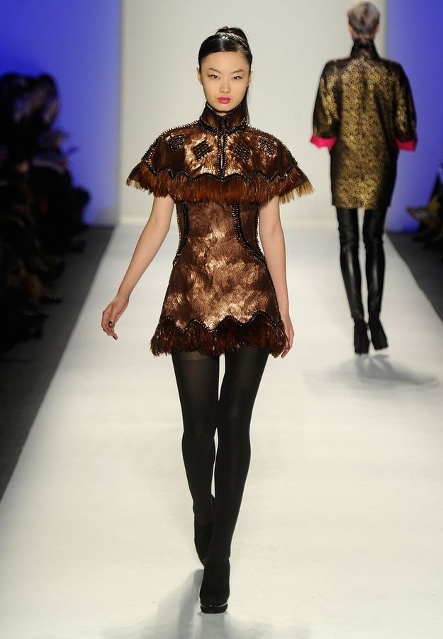 a woman in a brown dress and black tights walks down a runway during a fashion show.  she is wearing a black highheeled shoe.  her hair is pulled back and she has her hands by her side.  the background is blurry and other models can be seen.