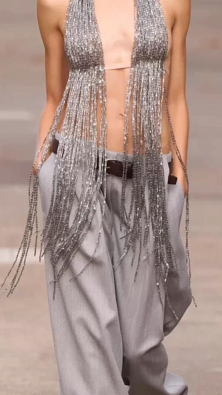 a fashion model poses on a runway, wearing a unique ensemble. she is dressed in highwaisted, light grey trousers that feature a dark brown leather belt with a prominent buckle. the top consists of a halterstyle beaded garment in silver, adorned with long, cascading fringe. her midriff is exposed between the top and trousers. the model has her hands in her pockets.