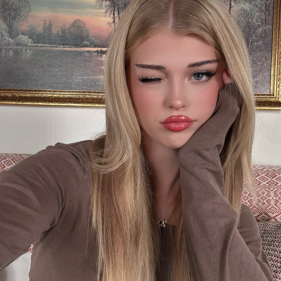 a young woman with long blonde hair is winking at the camera. she is wearing makeup and a brown long sleeve shirt with a necklace. behind her is a painting with a gold frame. she is sitting on a couch.
