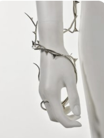 a closeup shot of a silver bracelet resembling thorns wrapped around a white mannequin hand against a white background. the bracelet features intricate details and a shiny finish, reflecting the studio lighting.