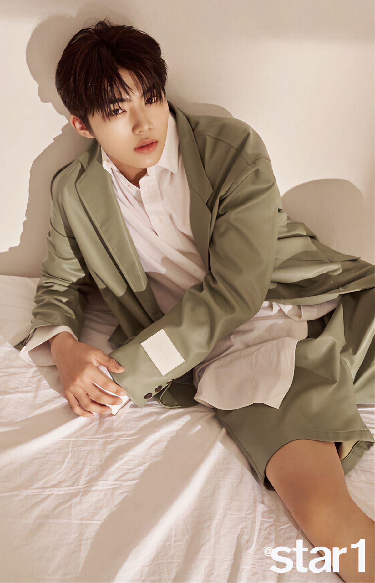 a young man is posing for a photoshoot. he is wearing a stylish green outfit, consisting of an oversized blazer and matching shorts.  he is sitting on a white bed with a relaxed posture. the lighting is soft and natural, creating a warm and inviting atmosphere.