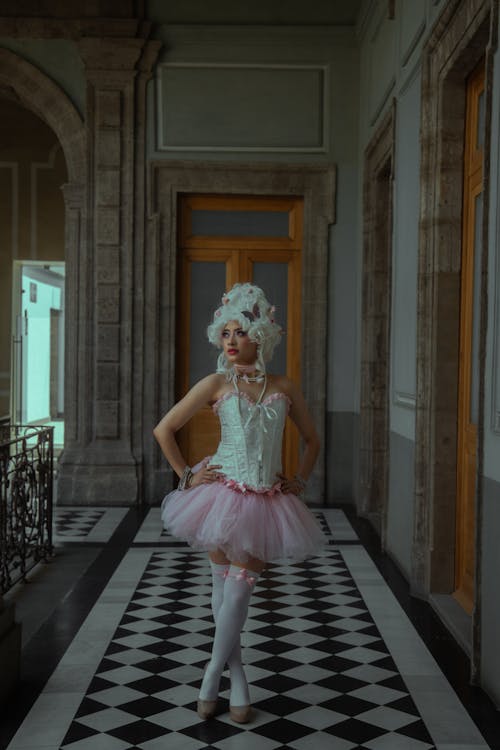 a woman in a pink and white tutu, corset, and large white wig stands in a long hallway with a black and white checkered floor. the architecture of the building is old and ornate. the woman looks elegant and graceful.